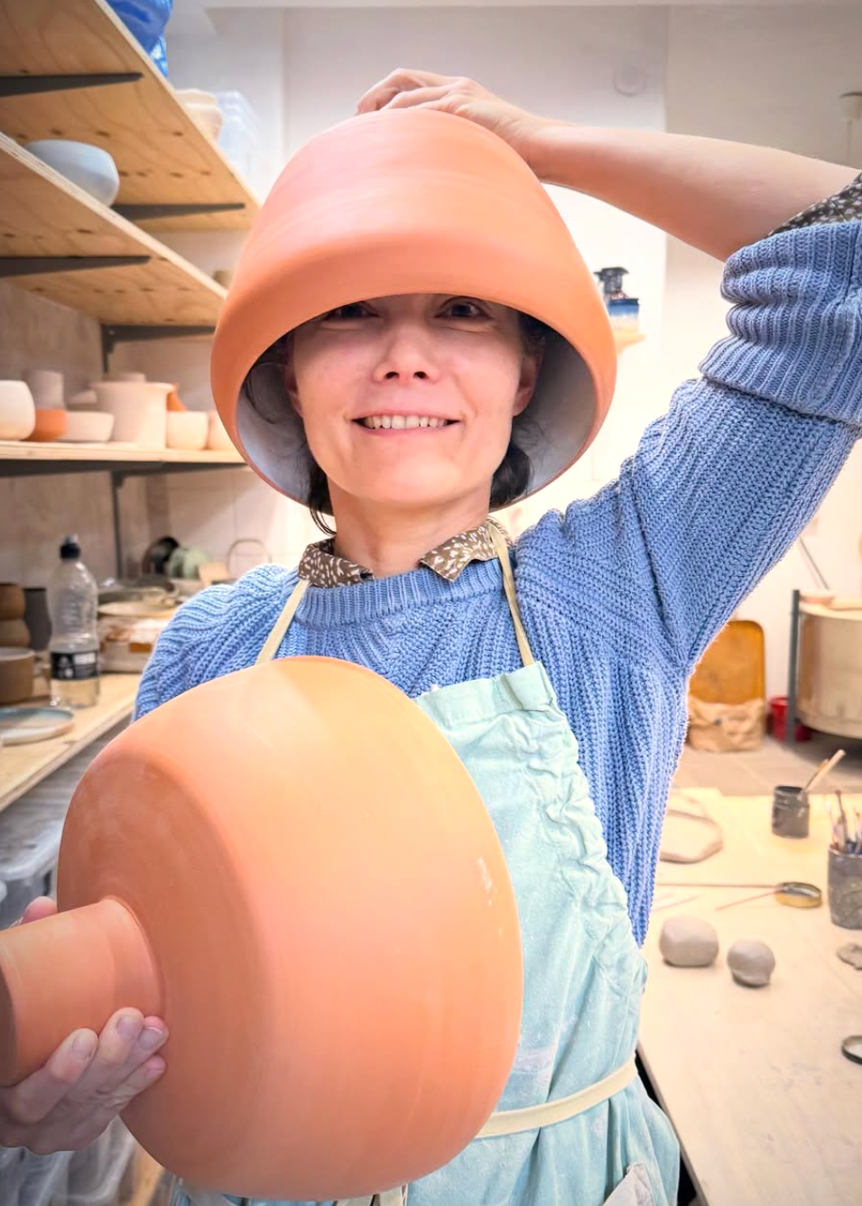 Person wearing a blue sweater and apron, holding a clay pot in a pottery studio.