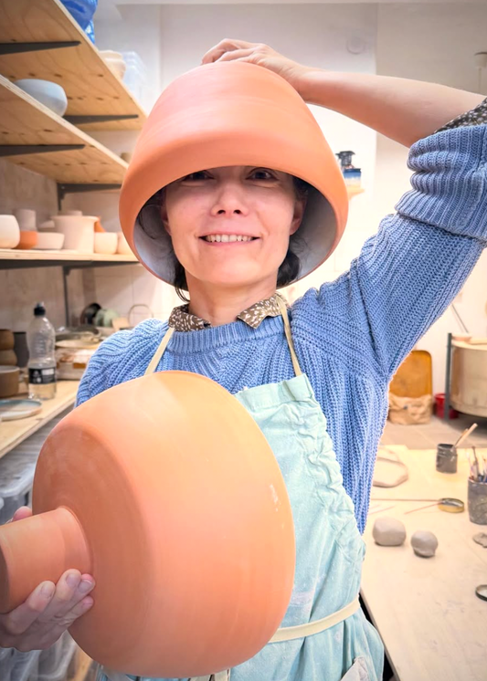 Person wearing a blue sweater and apron, holding a clay pot in a pottery studio.