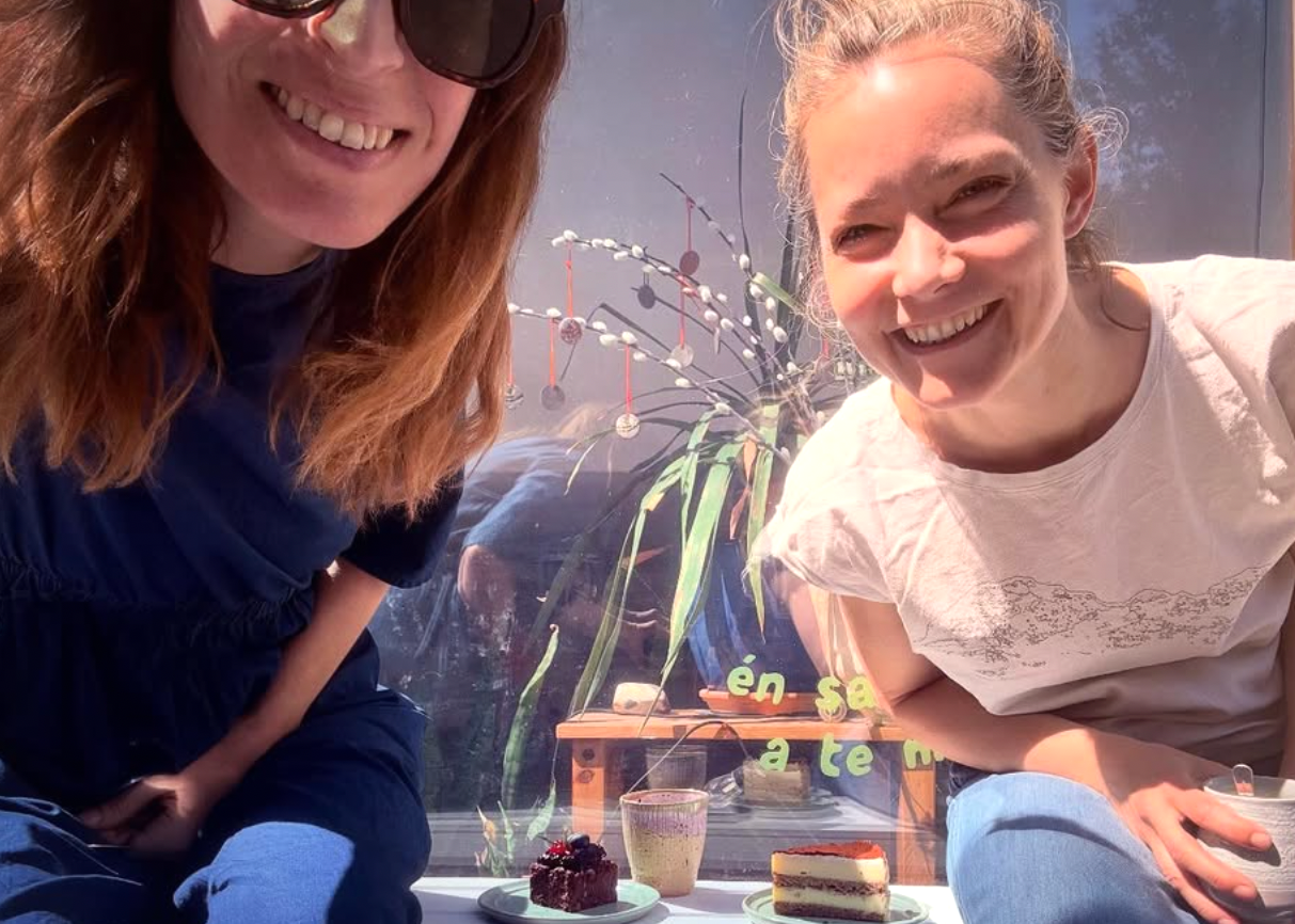 Two women sitting together, smiling, with a window and outdoor view in the background.