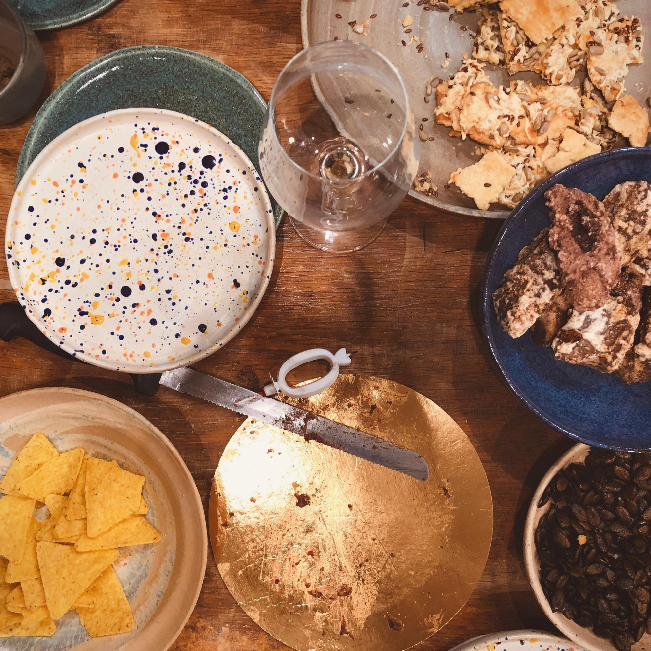 Diverse food items on a wooden table including tortilla chips, crackers, and a bowl of dark leafy greens.