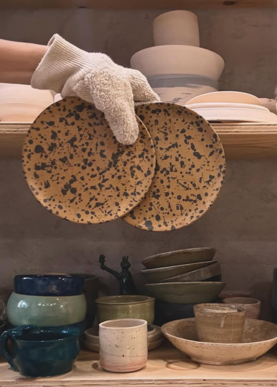 Person holding speckled ceramic plates with shelves of ceramicware in the background