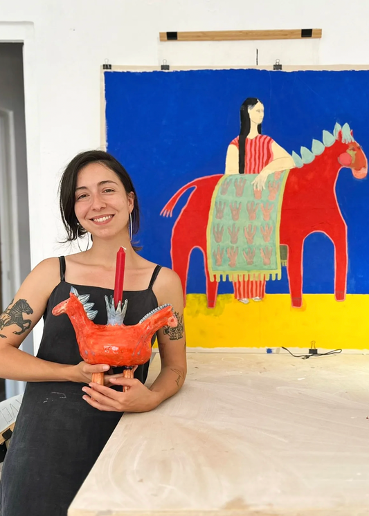 Woman holding a ceramic chicken in front of a colorful painting of a woman on a horse.