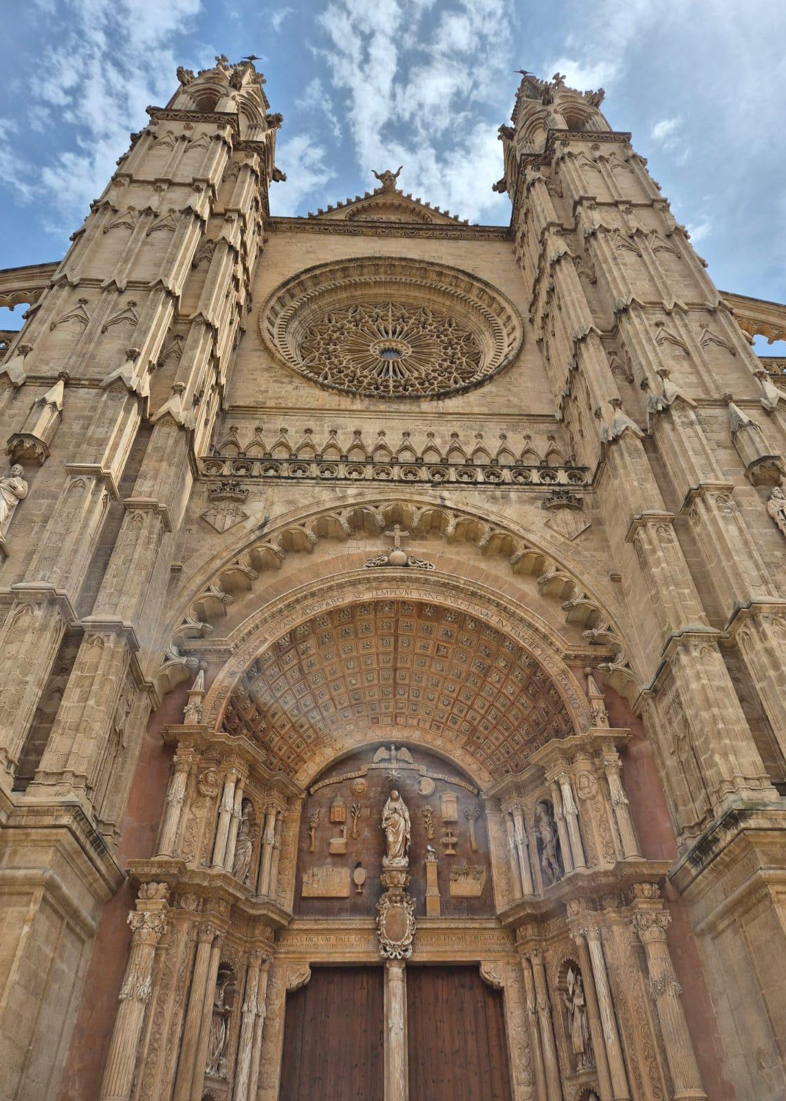 The grand facade of a Gothic cathedral, featuring ornate stone carvings, a large rose window, and a detailed central portal under a blue sky with light clouds.