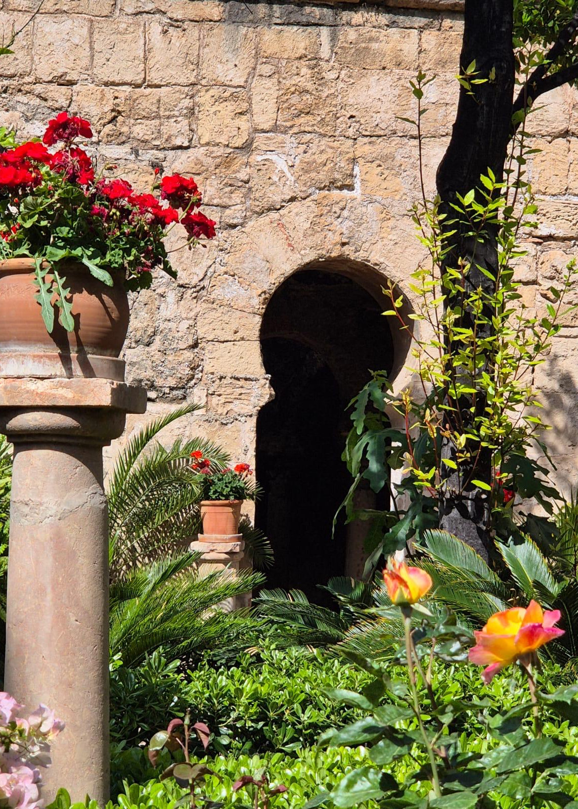 A lush garden courtyard with a stone wall, arched doorway, potted red flowers on a stone pedestal, and green foliage with orange and pink flowers in the foreground.