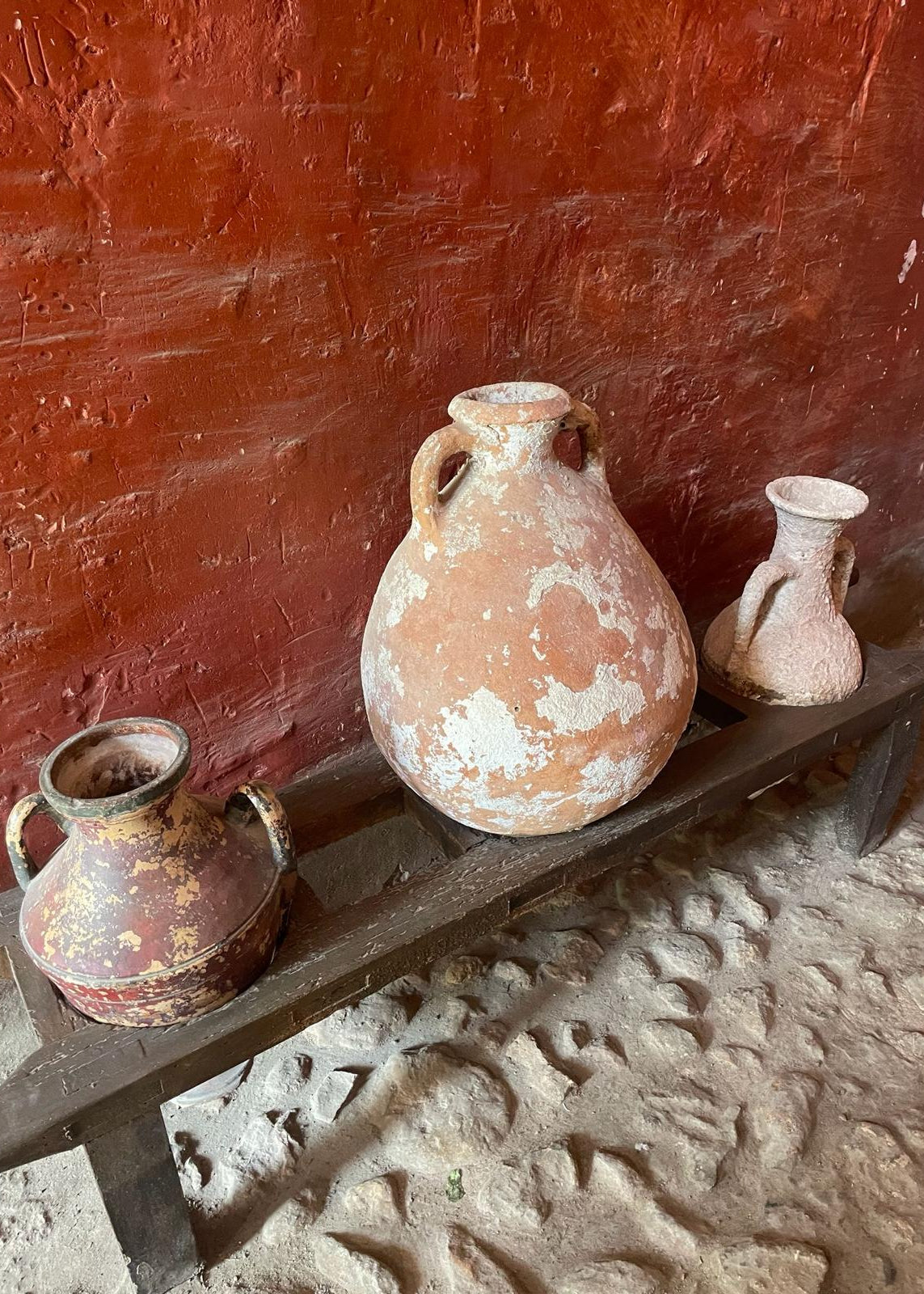 Three rustic terracotta clay jugs with handles displayed on a dark wooden bench against a textured reddish-brown wall and sandy floor.