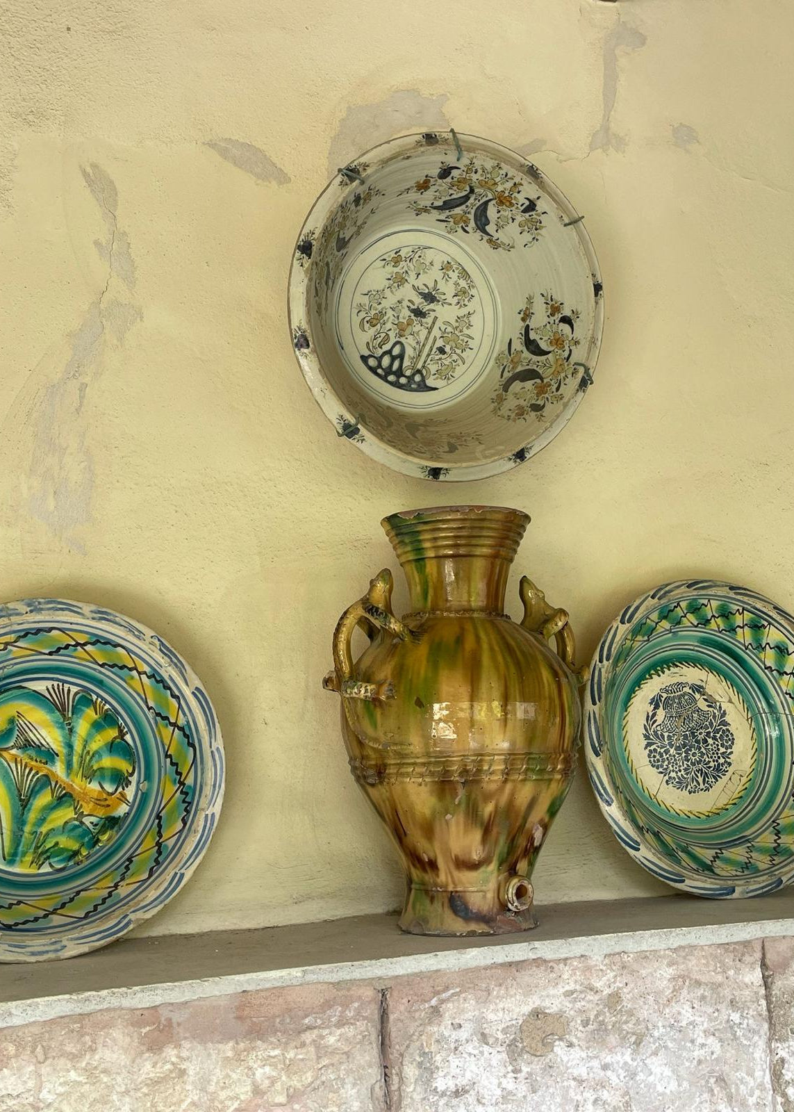 A rustic ceramic collection displayed on a stone shelf against a textured beige plaster wall, featuring a central green and brown glazed vase flanked by two patterned plates and a matching bowl above.