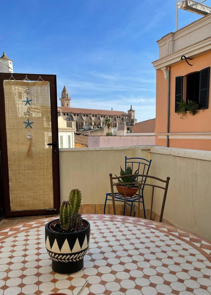 A balcony scene with a round table covered in a beige and white geometric tile pattern, a potted cactus in the foreground, a small chair, and a door with a woven panel. Beyond the railing, a historic building with towers is visible under a blue sky.