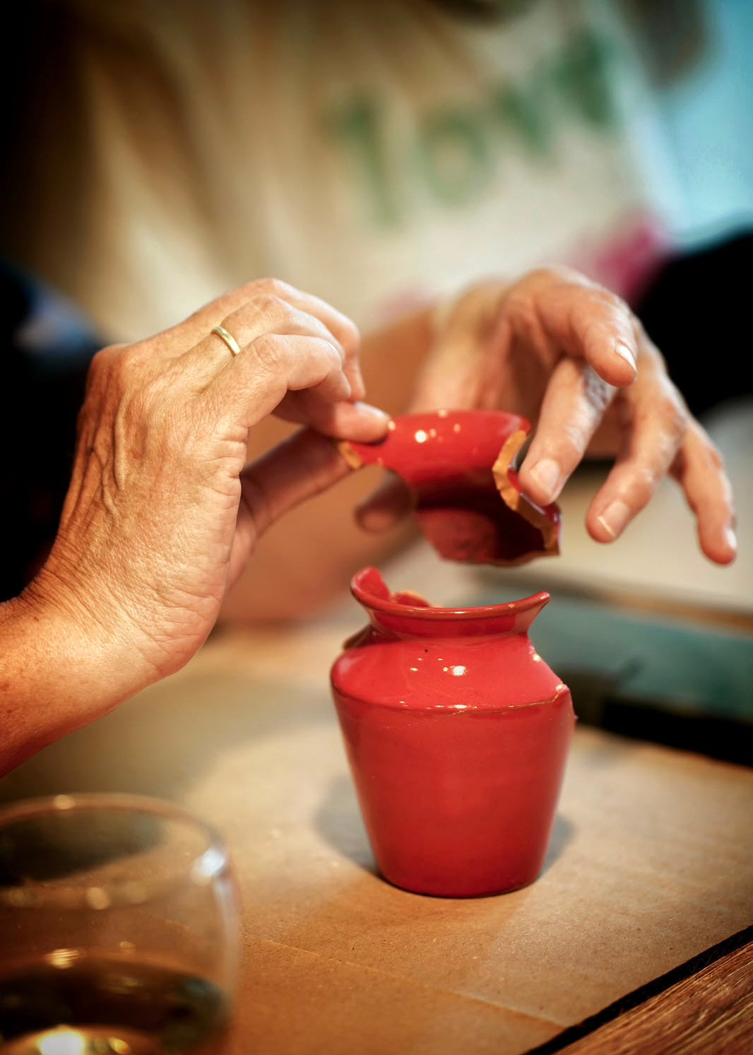 A close-up of a person's hands holding a small red ceramic cup above a matching red ceramic pot on a wooden table.