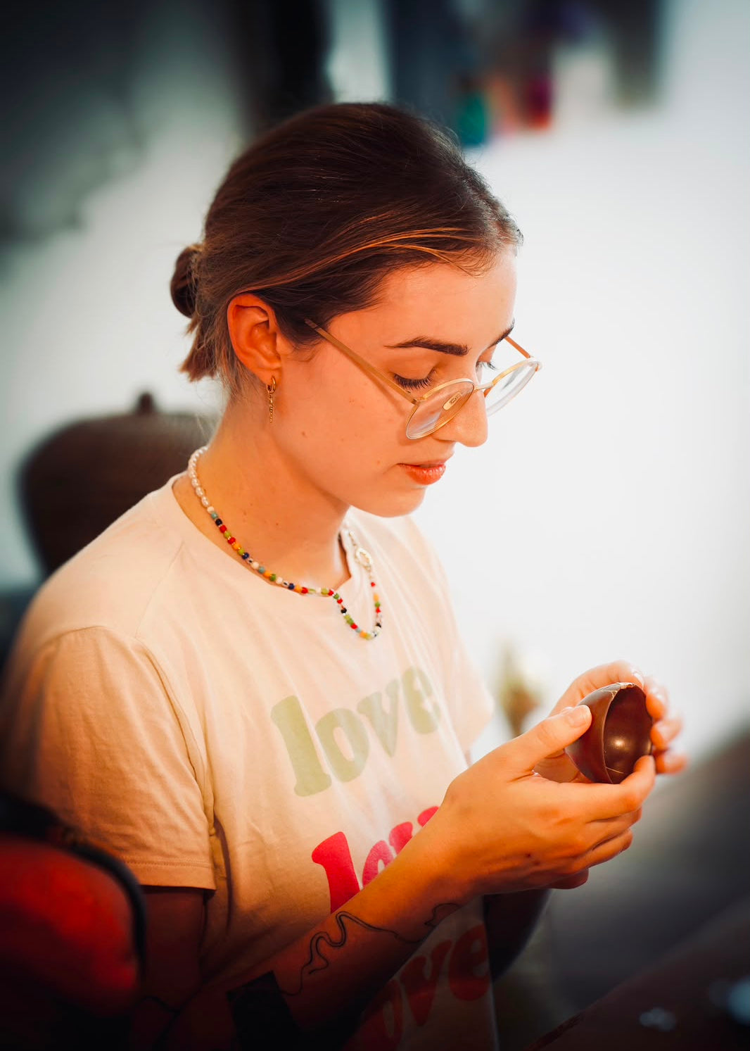A woman with glasses and a beaded necklace looks down while holding a small brown bowl in her hands, wearing a cream t-shirt with 'love' printed on it.