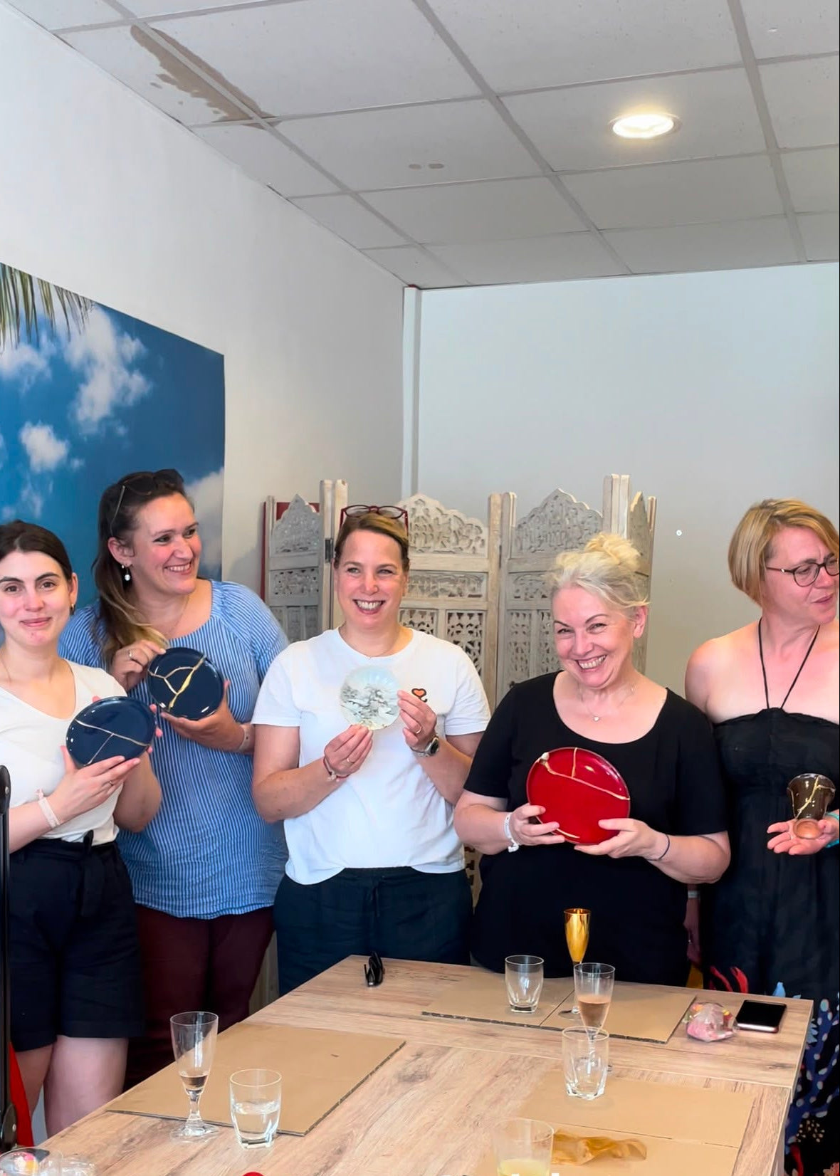 Five women standing together indoors, each holding a small round jewelry box or pouch in different colors including navy, black, white, red, and gold. They are gathered around a wooden table with glasses and cards on it, with a decorative screen and wall art in the background.