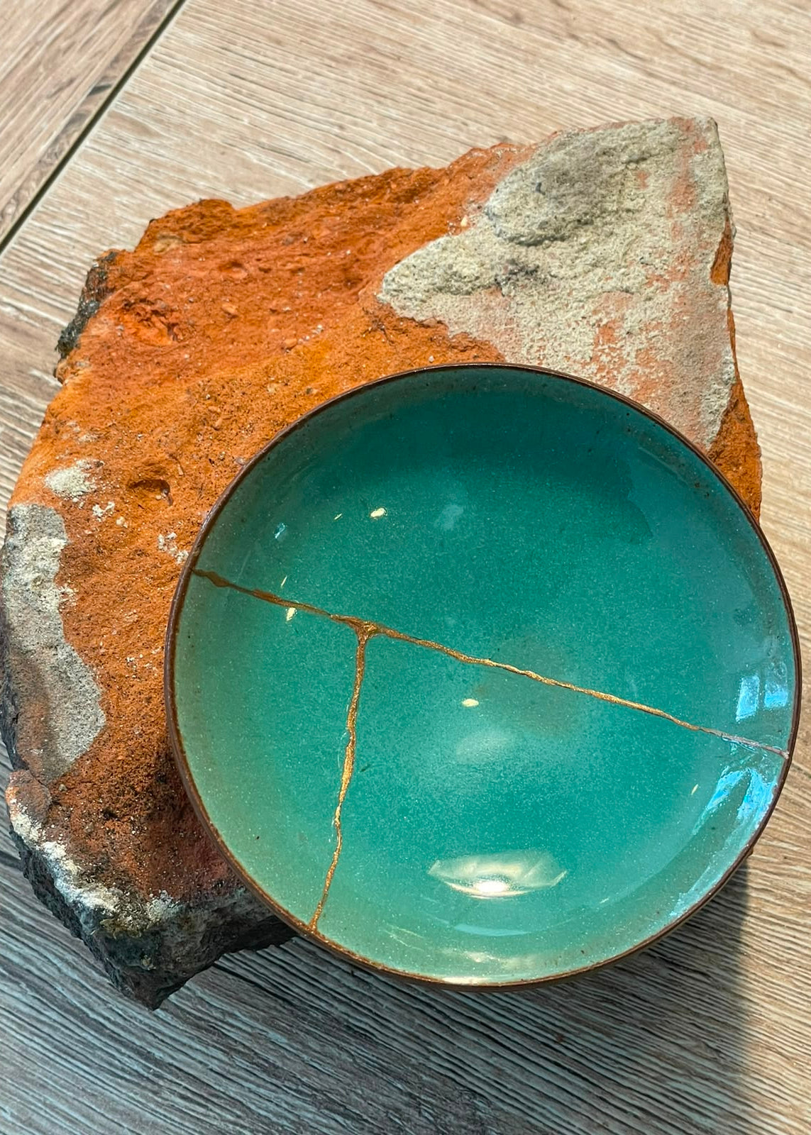 A handmade ceramic bowl with a glossy turquoise glaze and a visible gold repair crack, displayed on a textured orange and gray stone slab over a gray wood floor.