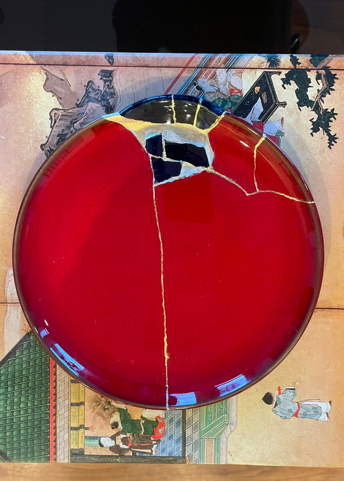 A round red ceramic plate with a glossy finish and a prominent gold crackle finish, featuring a large black repair patch and gold fissures on its surface, set against a traditional Japanese-style illustrated background.