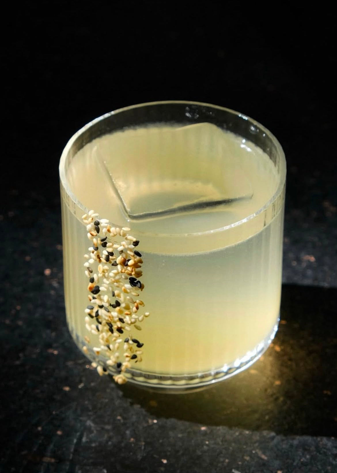A clear glass tumbler filled with a pale yellow liquid, photographed on a dark surface against a black background. A few small black and beige seeds or grains are clustered along the lower left side of the glass.