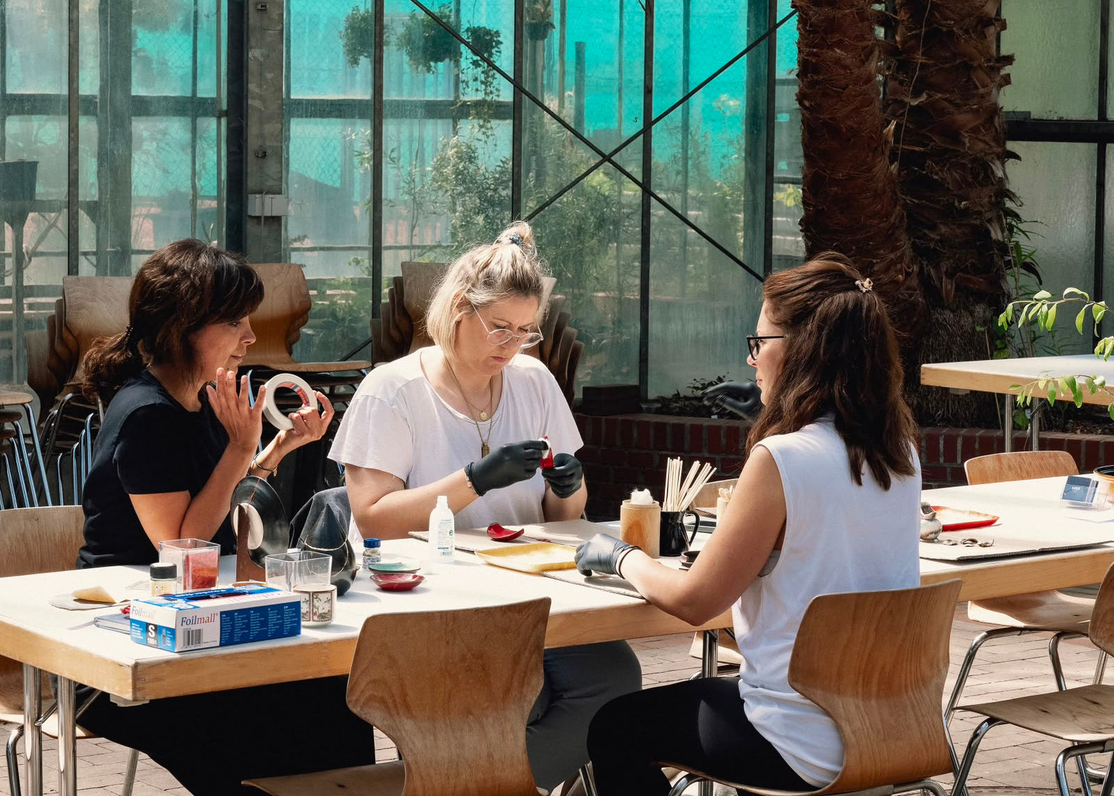 Three women seated at a wooden table in a bright indoor space with large windows and greenery, working together on craft or craft-related tasks with gloves on a table.