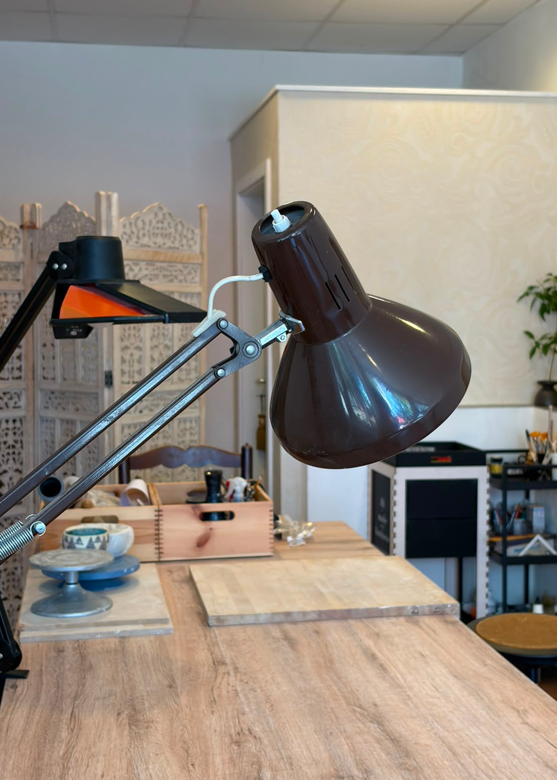 A dark brown adjustable desk lamp with a silver metal arm and black base, positioned on a wooden table in a modern office or studio setting.