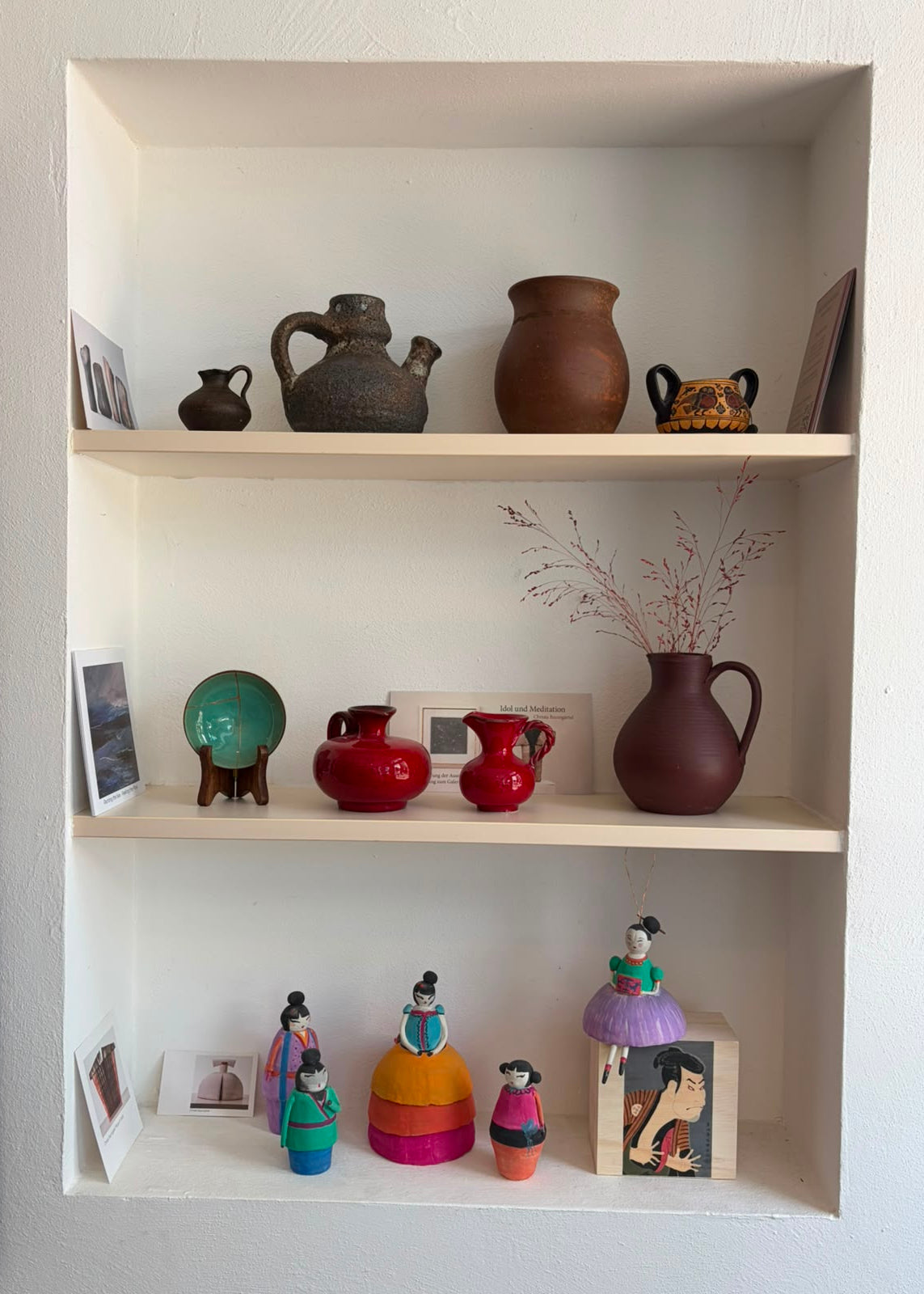 A set of three light wood shelves mounted on a white wall, displaying an assortment of decorative home items including ceramic pitchers, small figurines, and framed art.