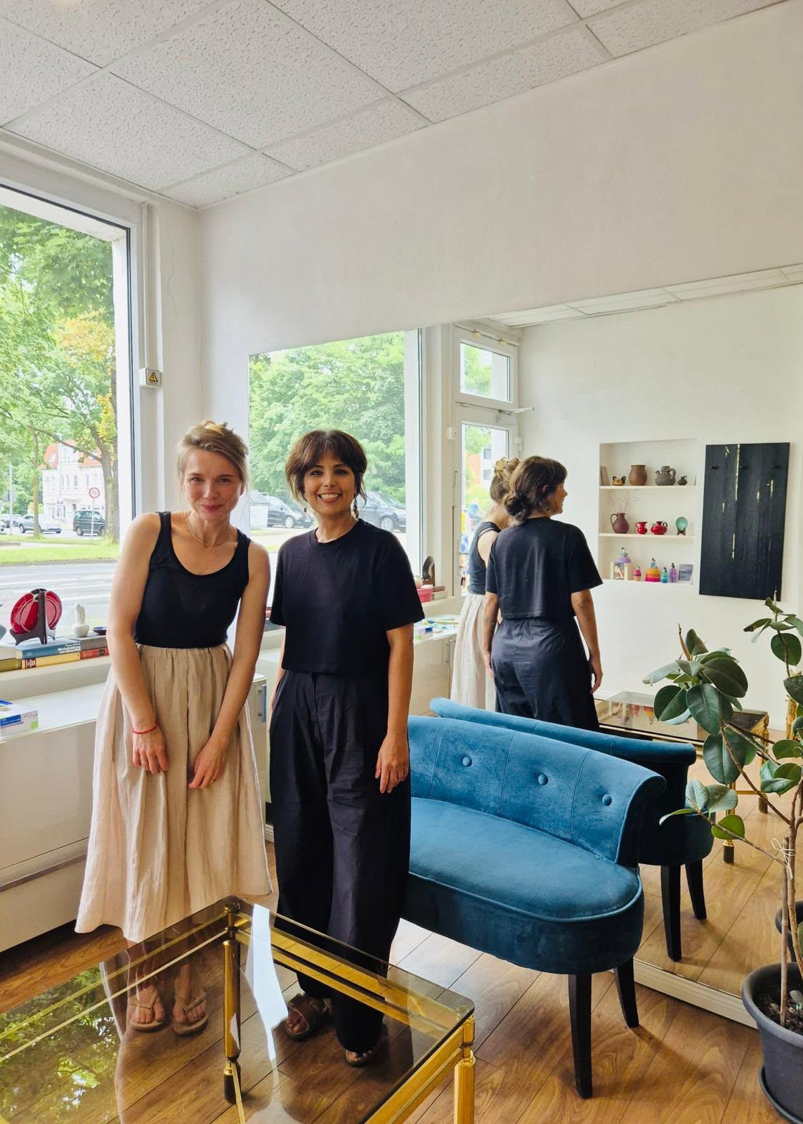 Two women posing in a bright, modern room with large windows, a blue velvet bench, and a mirror reflecting their back. One is wearing a black tank top and beige skirt, and the other is wearing a black t-shirt and dark trousers.