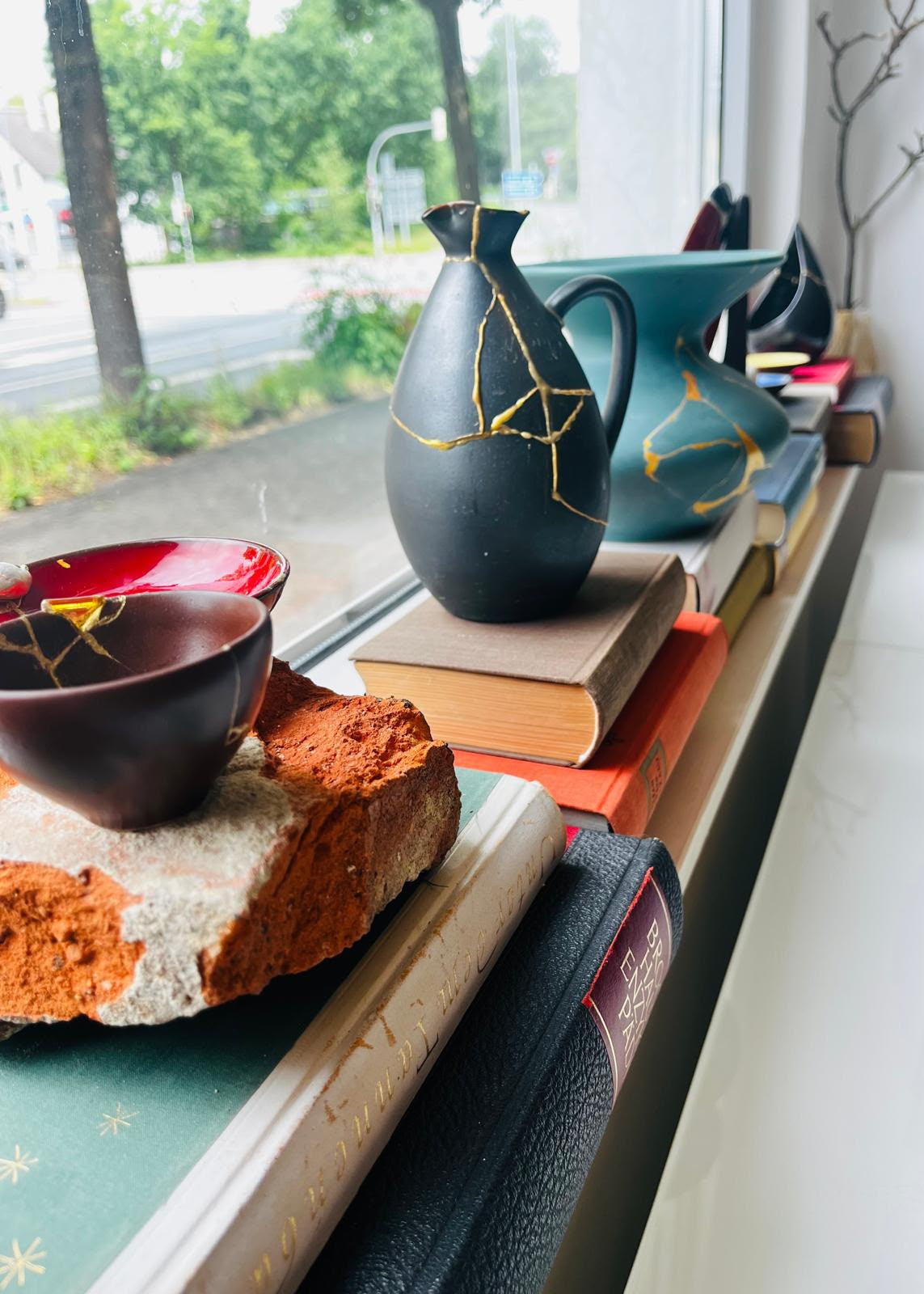 A collection of handmade ceramic vessels, including a black pitcher and bowl with gold crackle repair patterns, a teal vase, and a dark red bowl, styled on books on a windowsill with a real outdoor environment visible in the background.