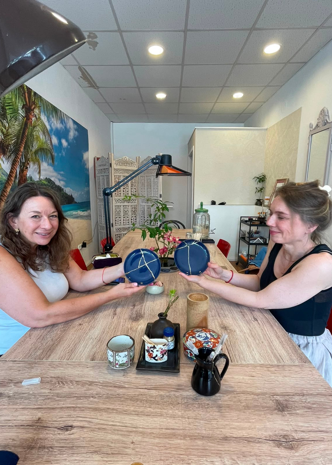Two women sitting across from each other at a wooden table, holding up round blue and white ceramic plates or coasters that feature a geometric pattern.