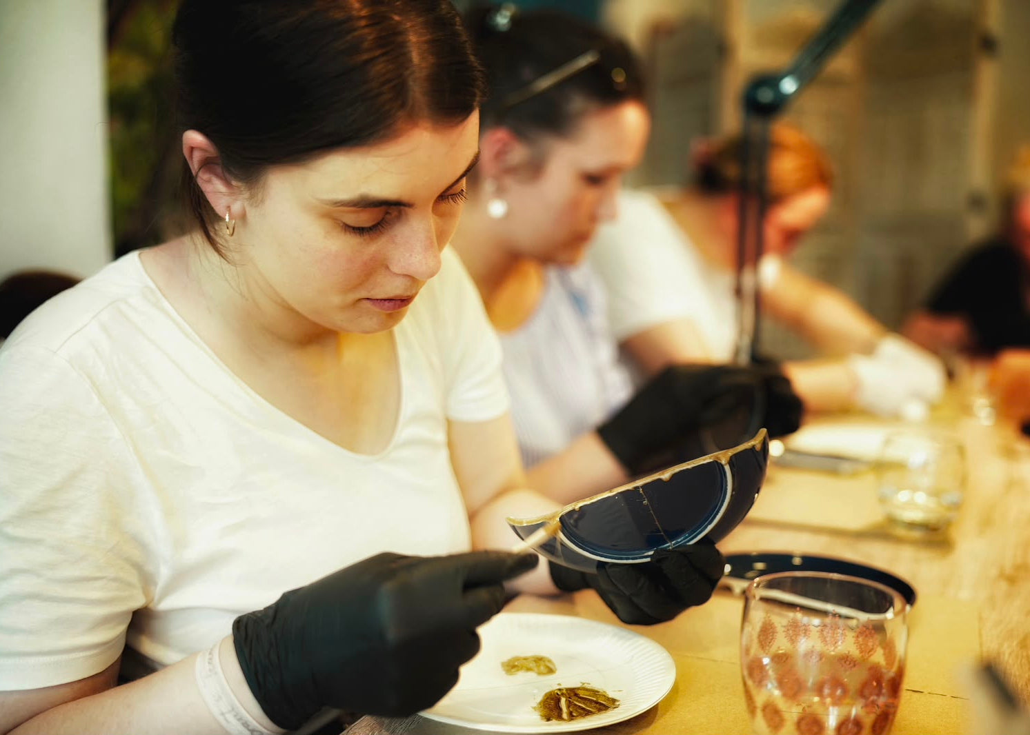 A close-up lifestyle shot of a woman in a white t-shirt and black gloves holding a pair of sunglasses with a black frame and blue lenses, working at a table with other people in the background.