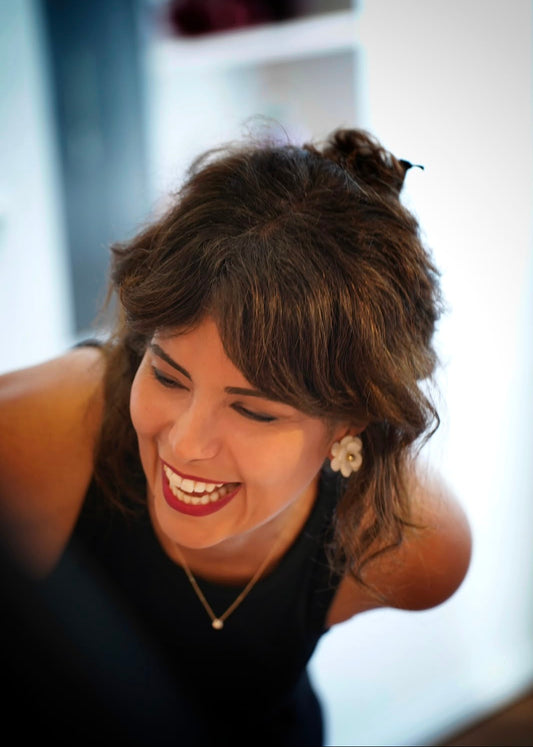 A smiling woman with brown hair in a half-up bun, wearing a black top and a delicate gold necklace, photographed in a brightly lit indoor setting.